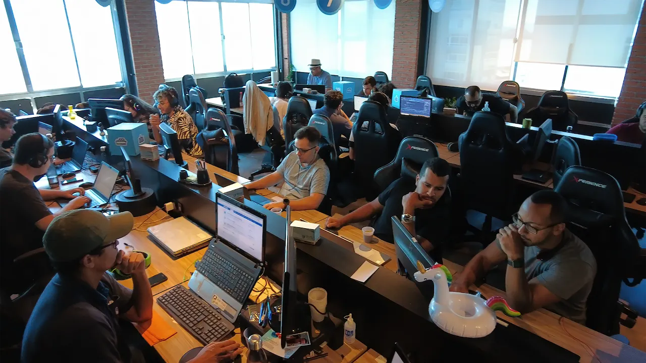 Escritório open-space da T2S com paredes de tijolos aparentes, desenvolvedores em cadeiras gamer e um unicórnio inflável sobre a mesa.